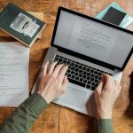 High-angle view of hands typing on a laptop surrounded by books and papers.